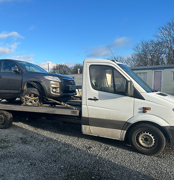 towing truck with a black SUV on a flatbed in a gravel area with blue sky and trees background four wheel drive vehicle transport